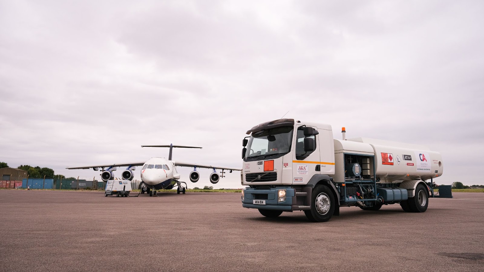A white aircraft refuelling vehicle for sustainable fuel parked on tarmac in front of a large blue and white research aircraft. The Cranfield Airport logo is visible on the side of the vehicle.